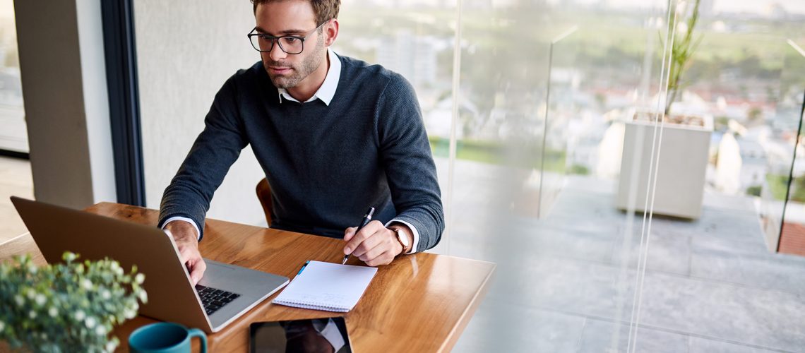 Young businessman sitting at a table at home working on a laptop and writing down ideas in a notebook